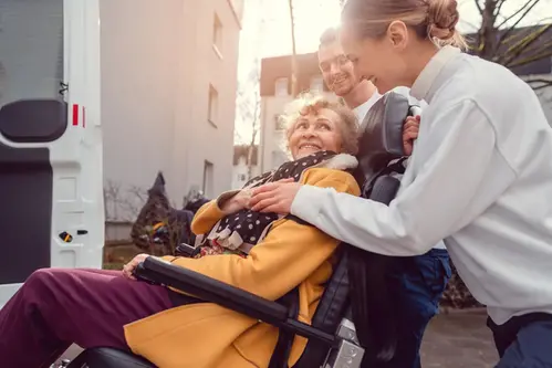 Kvinna i rullstol får hjälp upp i bil av annan person