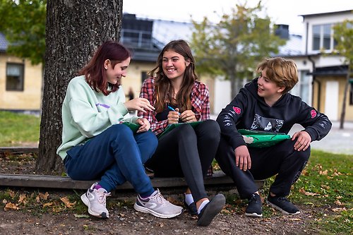 Tre ungdomar sitter utomhus i gräset och skrattar med varandra