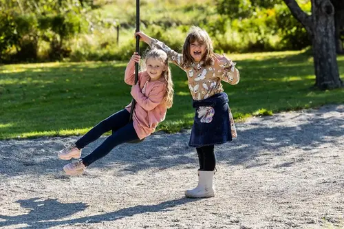 Två glada flickor leker på solig skolgård med linbana